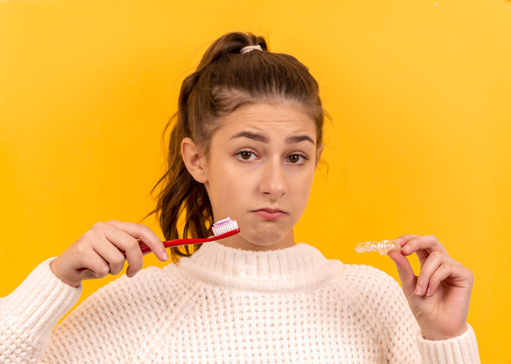 A teenage girl with a ponytail and a white sweater holding a toothbrush and a clear aligner confused